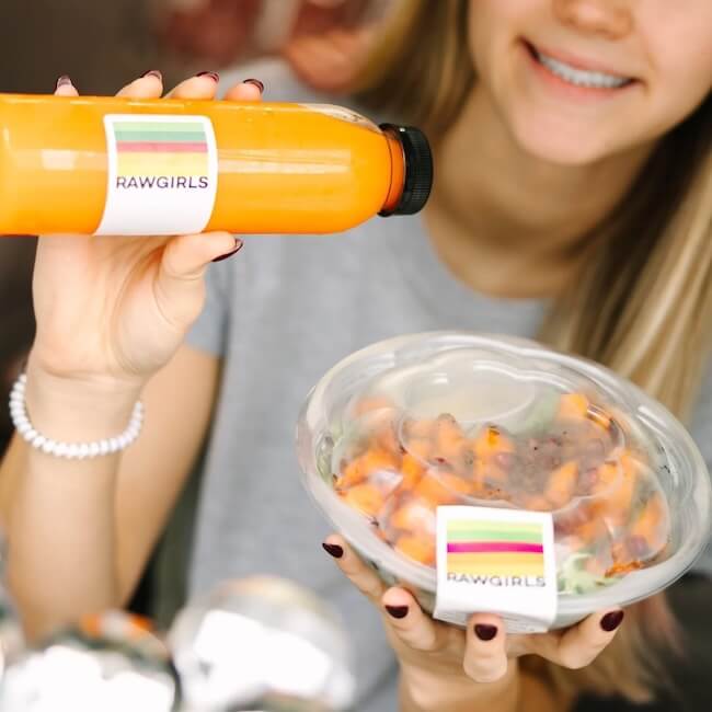 Person holding an orange juice bottle and a bowl of fruit, both labeled "RAWGIRLS," a spot known for the best salads in Memphis.