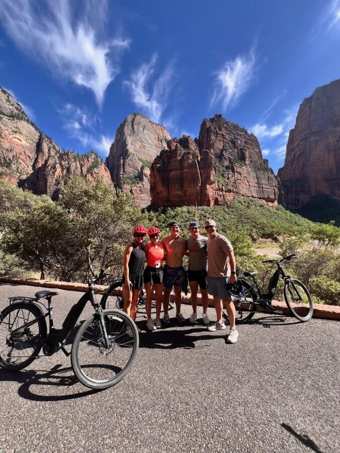 Meredith Mann and her adventurous crew of five cyclists pause on the road, their bicycles framed by rocky mountains and a brilliant blue sky.