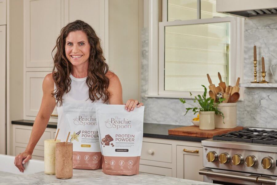 Meredith Mann stands in a kitchen next to two bags of protein powder labeled "The Peachie Spoon," with two smoothies on the counter.