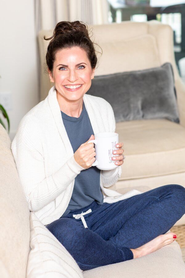 Meredith Mann smiles while sitting on a couch, holding a mug. She’s wearing a white cardigan and blue lounge pants, enjoying her peaceful morning.