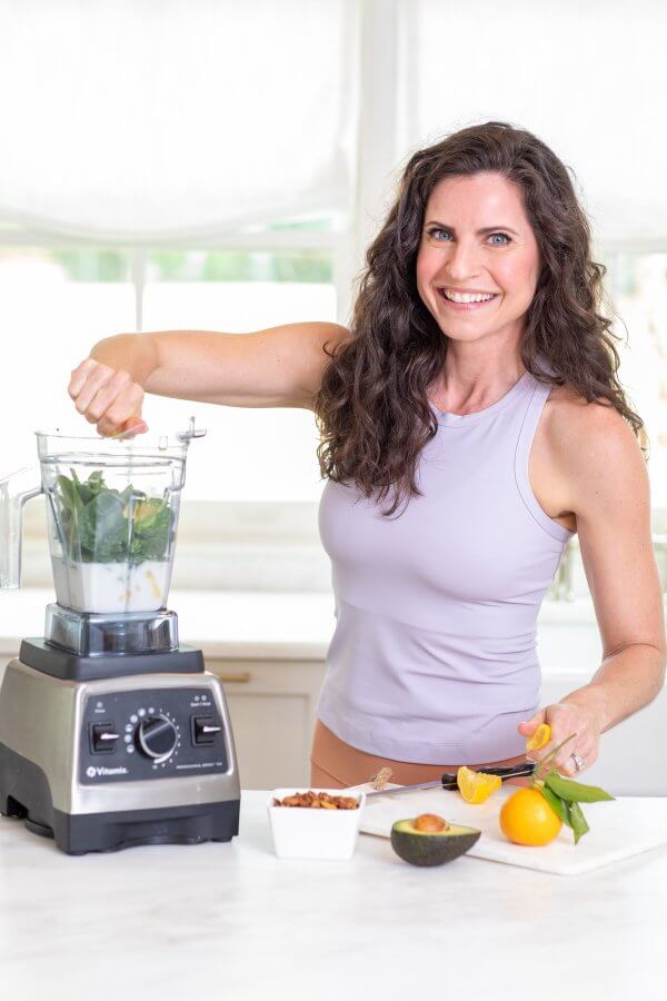 Meredith Mann, in a sleeveless top, skillfully prepares a smoothie in her blender, blending spinach, an orange, almonds, and avocado amidst the cozy ambiance of her kitchen.