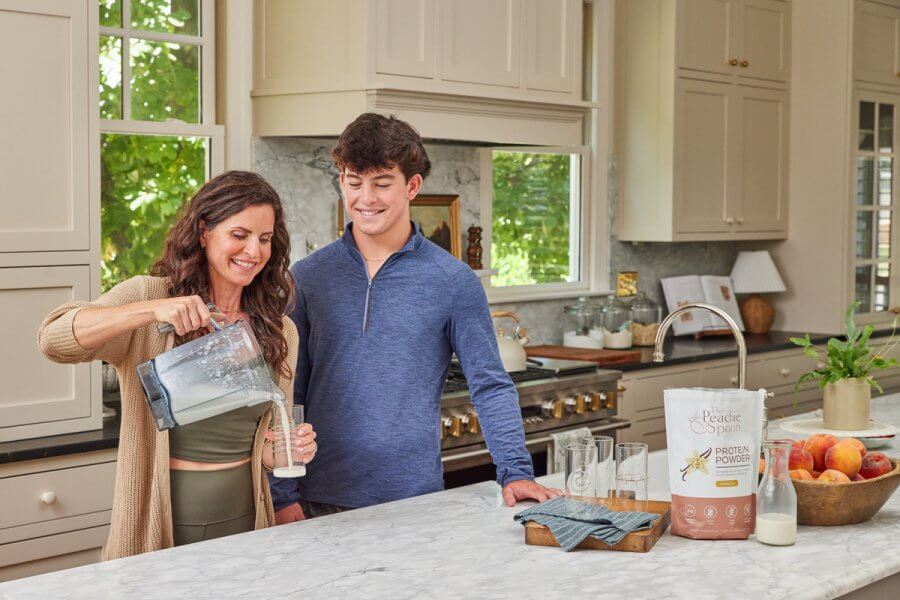 In the kitchen, Meredith Mann pours a smoothie into a glass, standing beside a young man. On the counter lie a bag of Peachie Spoon protein powder and a selection of fruits.