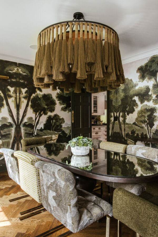 Dining room featuring a large round table with patterned chairs, a unique chandelier with hanging fabric elements, and walls adorned with a forest-themed mural, showcasing exquisite design influenced by Atlanta's vibrant aesthetic.