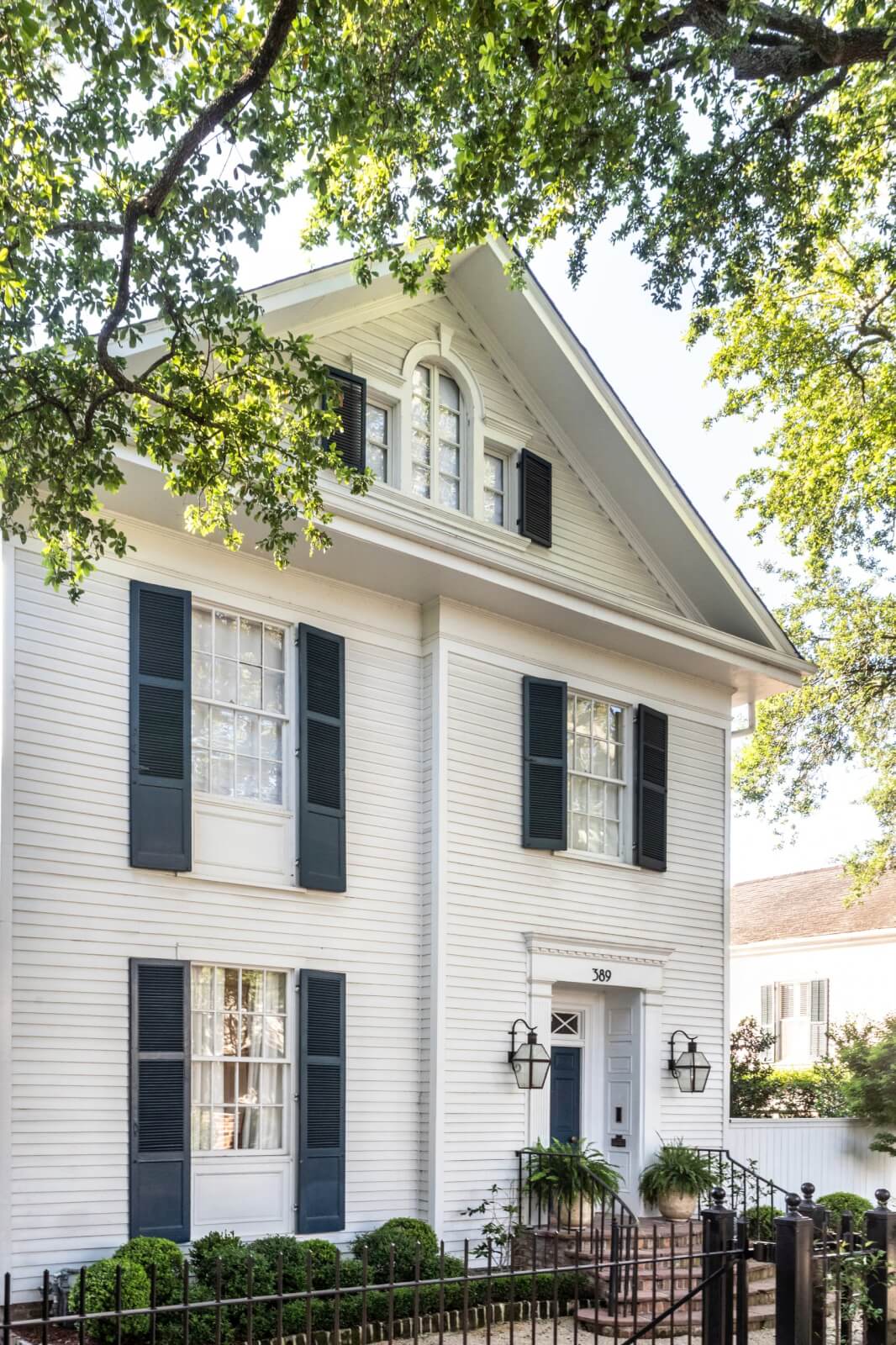 A white 1800s home in New Orleans with black shutters, arched windows, and a small front garden is surrounded by a black fence under leafy trees. 