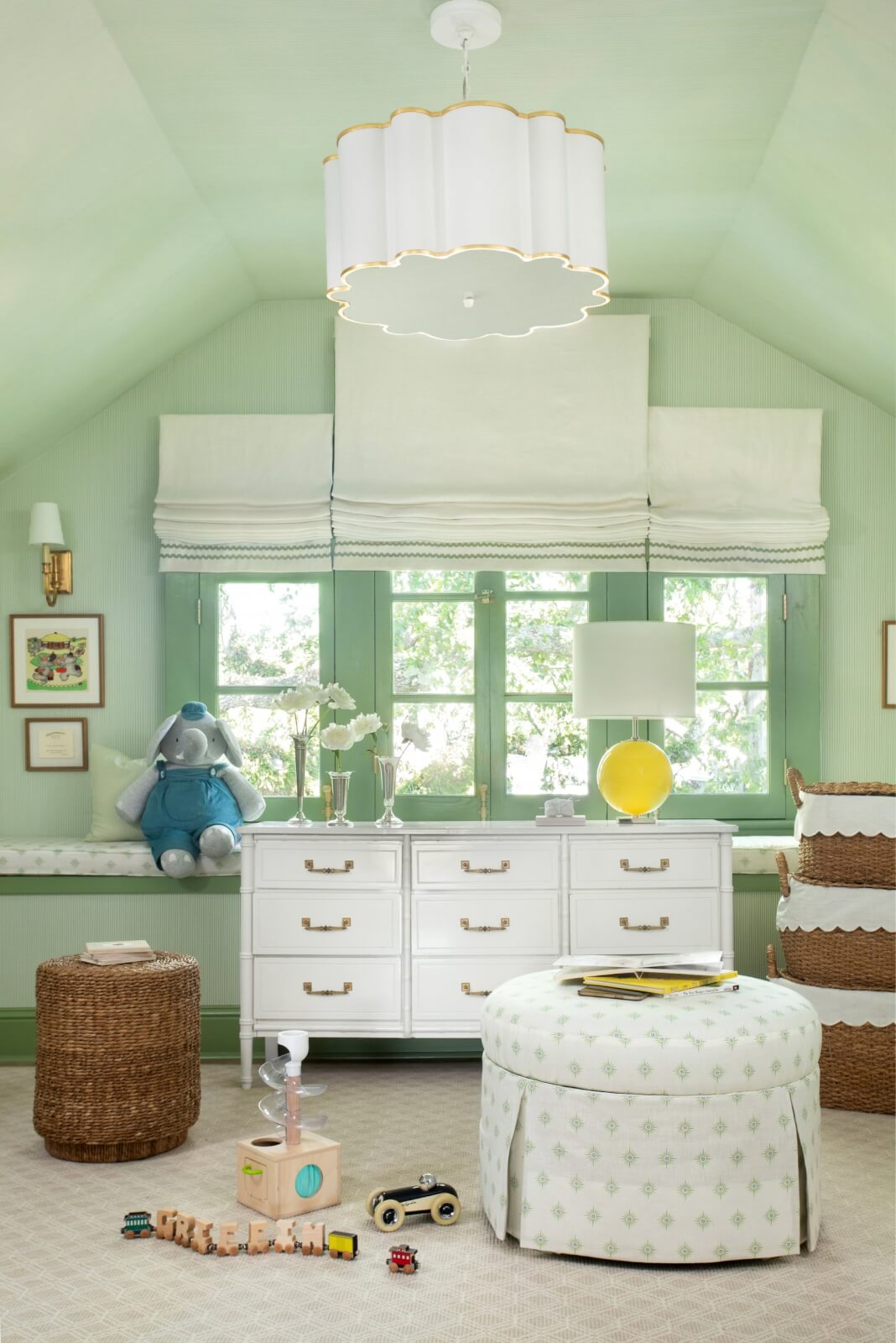 A pastel green nursery in a charming New Orleans home features a white dresser, plush toys, a round ottoman, and a woven basket. Large windows fill the room with natural light, while a cloud-shaped ceiling light hangs above.