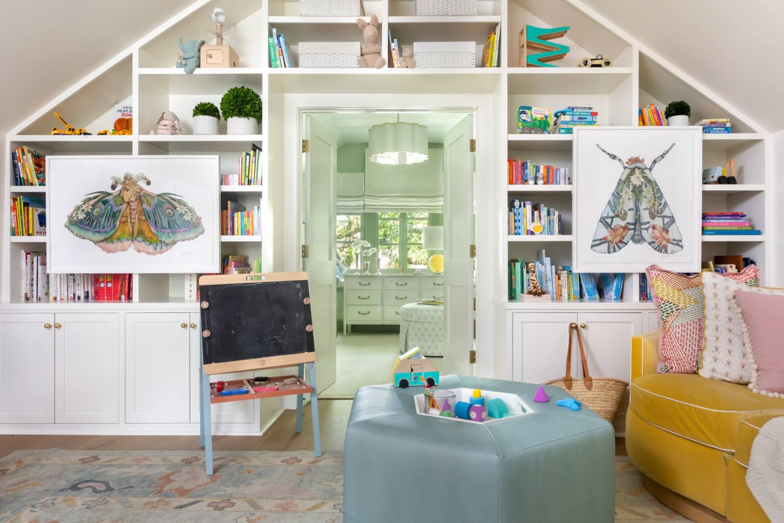 White built-in bookshelves filled with books and decor bring charm to the colorful children's playroom, accentuated by two framed butterfly artworks. A chalkboard and a teal ottoman with toys add playful touches. 