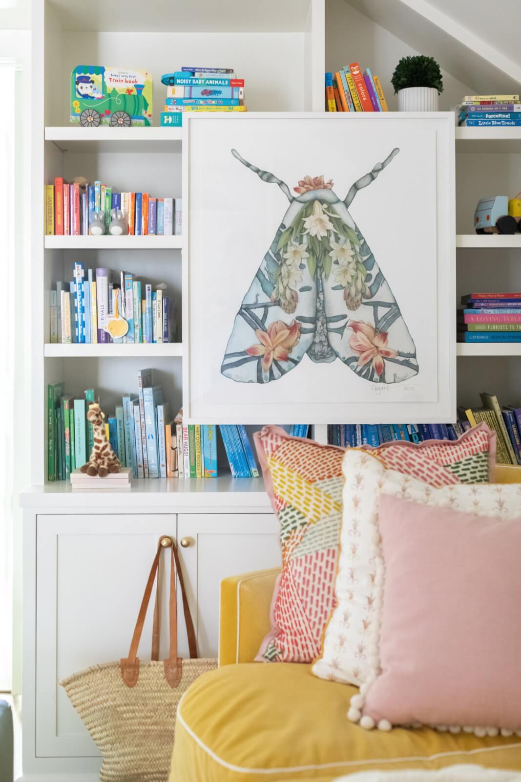 A cozy, colorful New Orleans home reading nook features a cushioned yellow sofa with patterned pillows, a decorative moth print, and a variety of books on the white shelves behind.