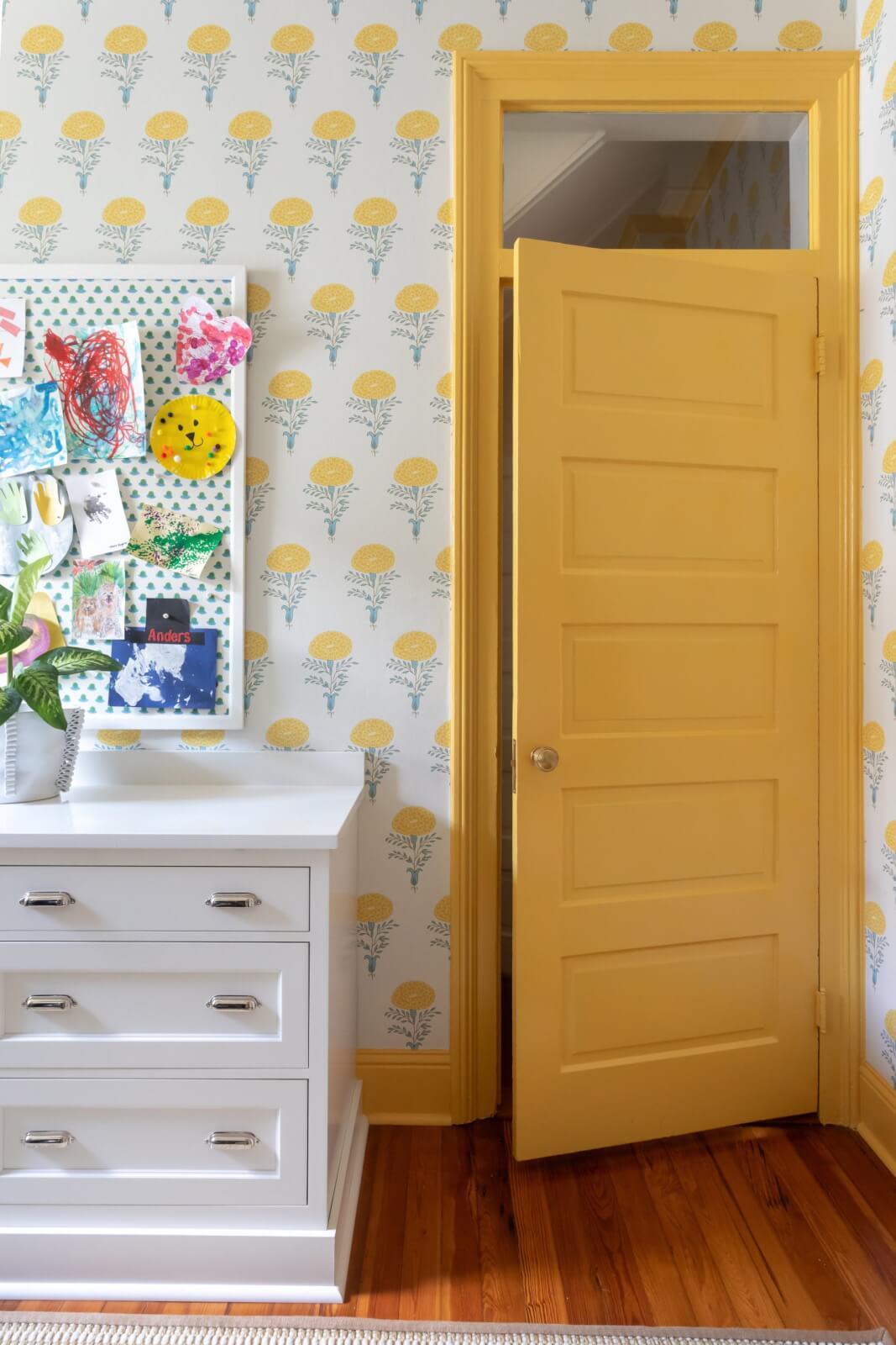 Room with a yellow door, white dresser, colorful children's artwork, and pretty floral wallpaper.