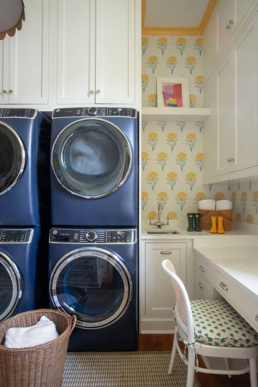 Laundry room in a New Orleans home featuring stacked blue washers and dryers, white cabinets, and a small sink. A chair sits beneath the pretty wallpaper in yellow and blue hues, adorned with a shelf showcasing a potted plant and artwork.