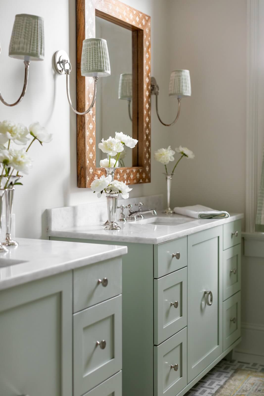 In a charming New Orleans home, the bathroom boasts light green cabinetry. A large mirror and two wall-mounted lamps enhance the space, while marble countertops showcase elegant white floral arrangements in vases.