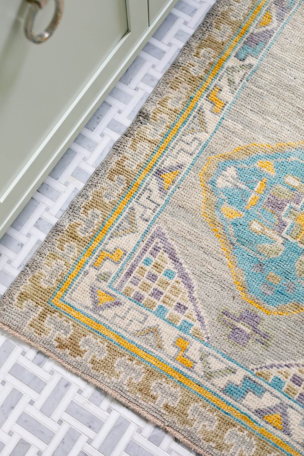 A patterned rug with geometric designs in blue, yellow, and gray graces a white woven floor near a light green cabinet.