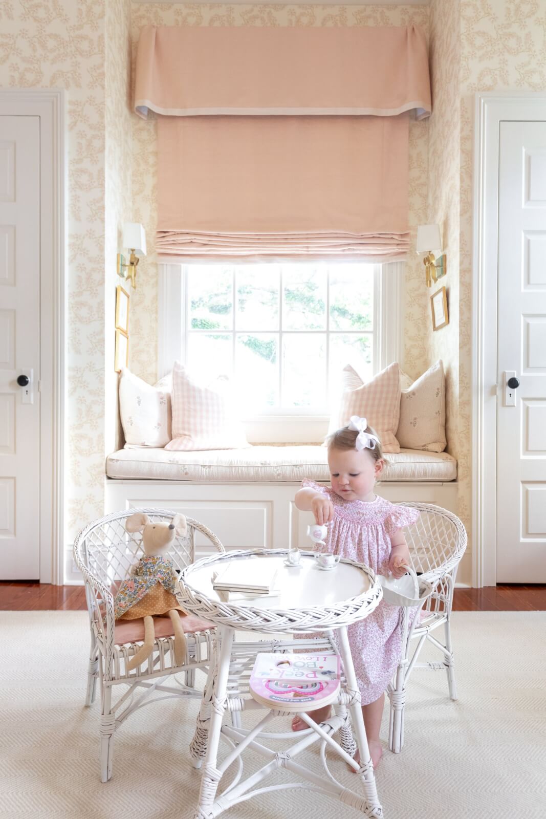 In a cozy New Orleans home, a child in a pink dress enjoys a tea party with a stuffed animal at a small white table. The room features soft decor and a charming window seat.