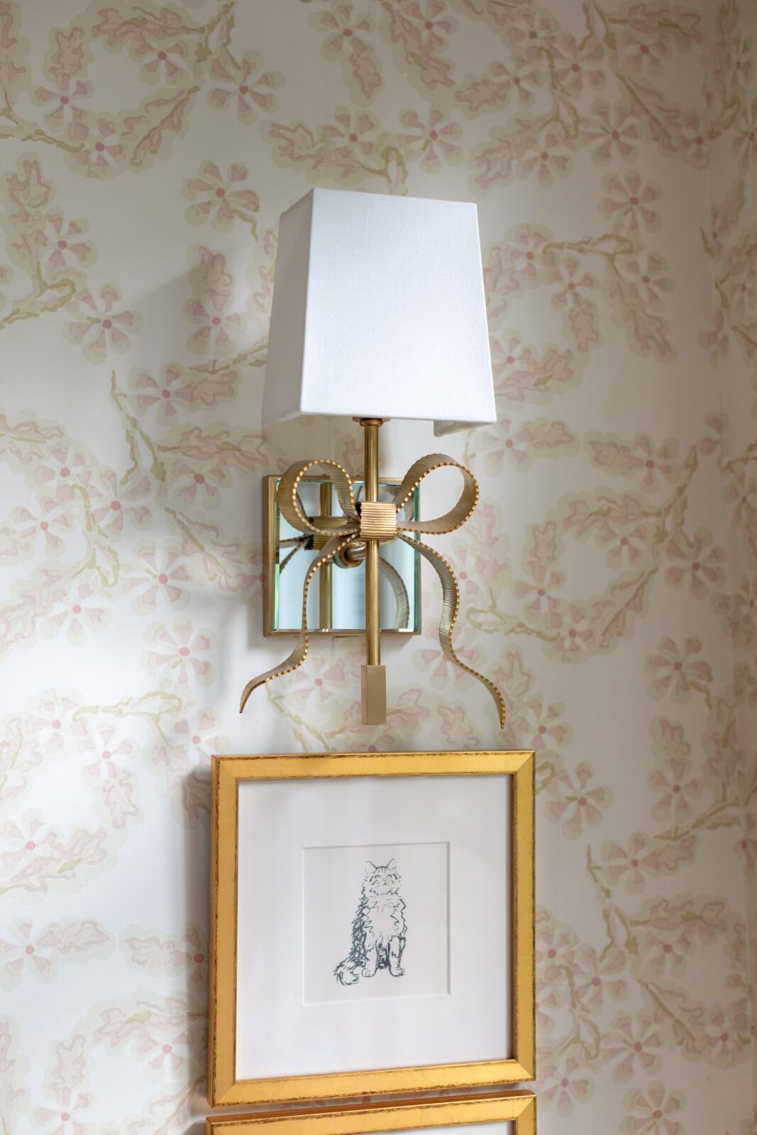 A decorative wall fixture with a white lampshade and bow design hangs above a framed sketch of a cat.