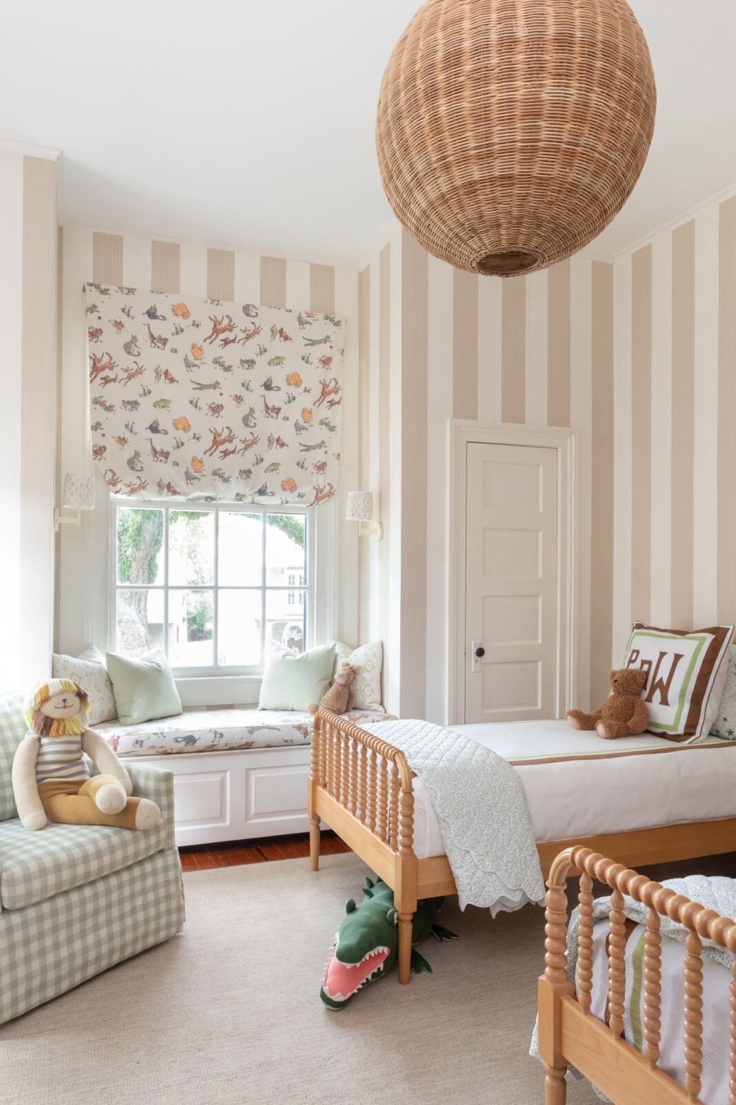 This cozy New Orleans home features a children's bedroom with pretty wallpaper adorning its striped walls. It includes a charming window seat, a single bed flanked by stuffed toys, and is illuminated by a wicker basket light fixture. A playful crocodile toy rests on the floor.