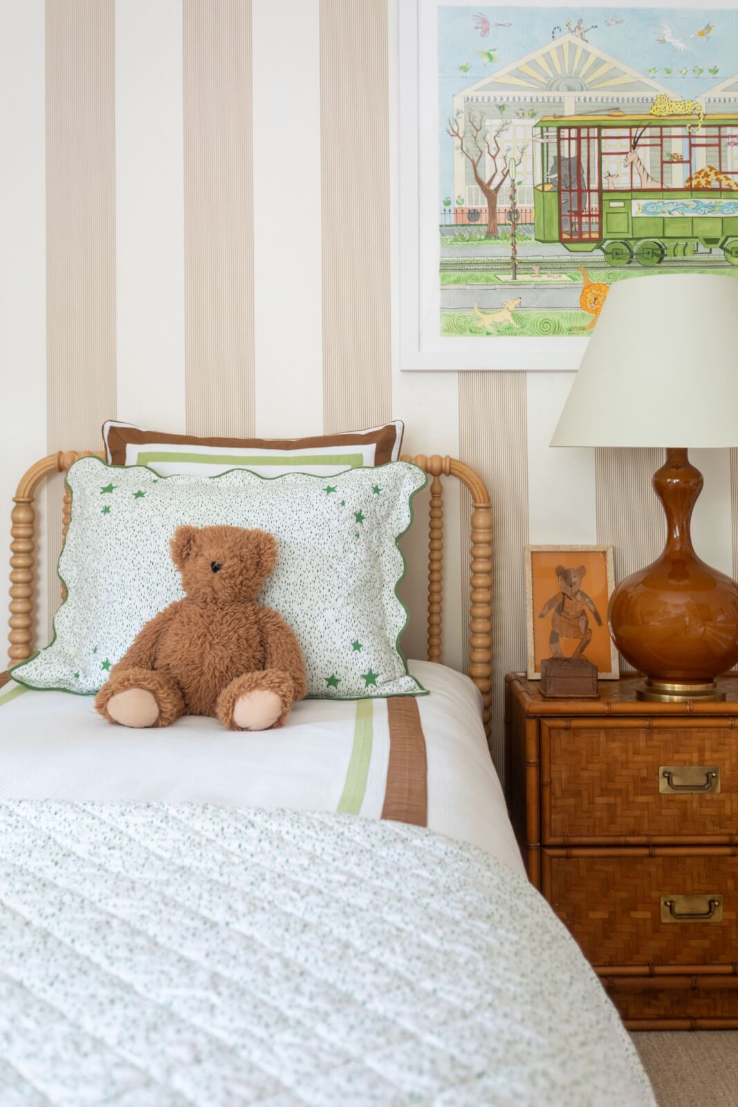 A cozy bedroom in a New Orleans home features a wooden bed, green and white bedding, and a teddy bear resting on the pillow. The room is accented by a brown lamp and framed artwork against the backdrop of pretty wallpaper, adding charm to this inviting space.