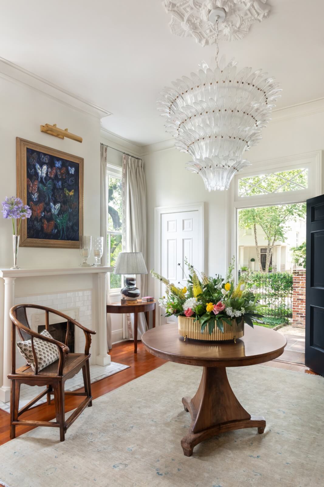 The elegant New Orleans foyer features a chandelier, round wooden table, floral arrangement, and painting above the fireplace. A classic wooden chair rests against tastefully patterned wallpaper, while an open black door offers a charming view of the garden beyond.