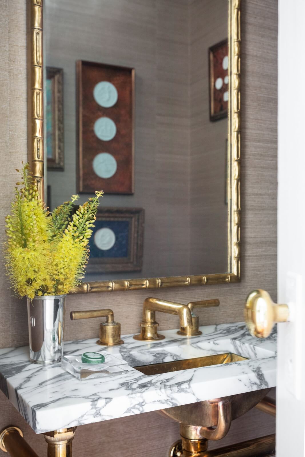 This New Orleans home bathroom features a marble countertop with gold fixtures and a vase of yellow flowers. A large mirror above the sink reflects framed art.