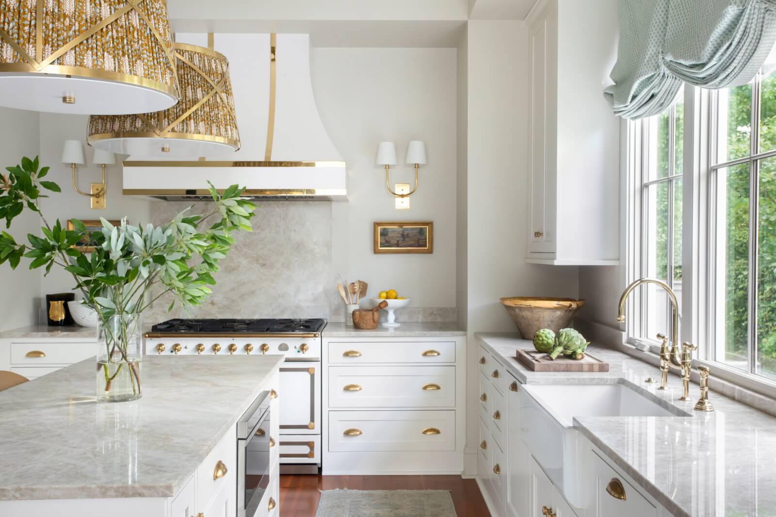 A bright kitchen with white cabinets, a marble island and countertops, gold accents, and hanging light fixtures exudes New Orleans charm. A potted plant adds a touch of nature, while elegant wallpaper complements the window's draped curtains.