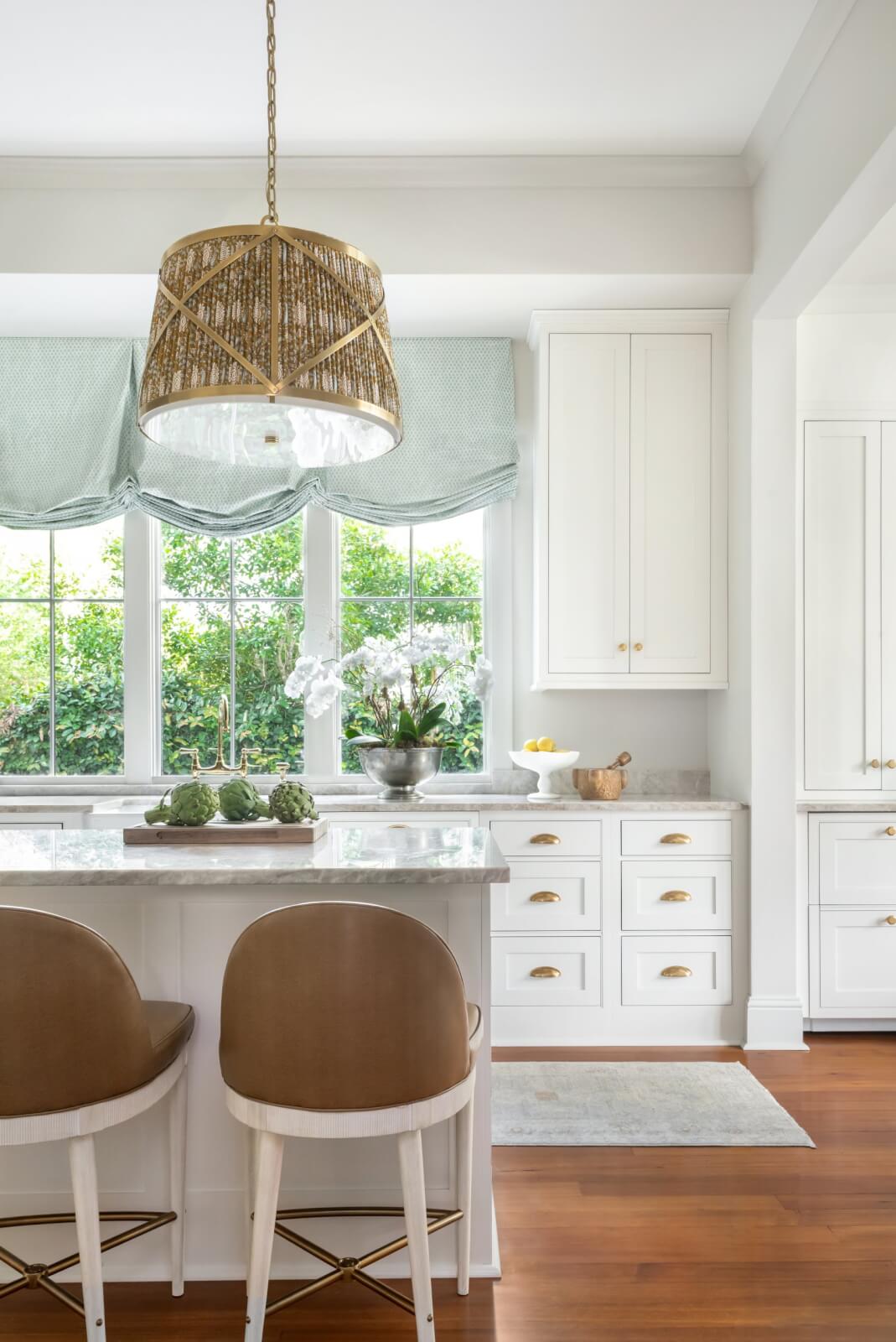 Bright kitchen with white cabinets and a marble countertop island, complemented by two brown stools and a wicker pendant light. The large windows with green curtains add charm to this inviting home.