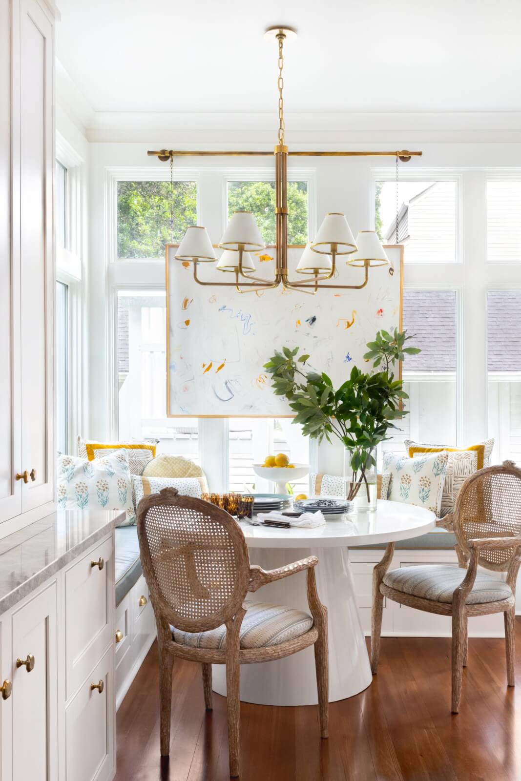 A bright dining nook in a New Orleans home features a round white table, wicker chairs, a built-in bench, and large windows. A chandelier hangs overhead. A decorative plant sits elegantly on the table.