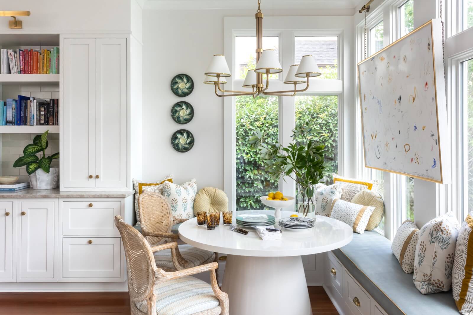 Bright kitchen nook with a white round table, cushioned bench, and wicker chairs. A large window opens to lush greenery, while built-in shelving displays decorative plates. A hanging abstract artwork complements the charming New Orleans-inspired wallpaper.