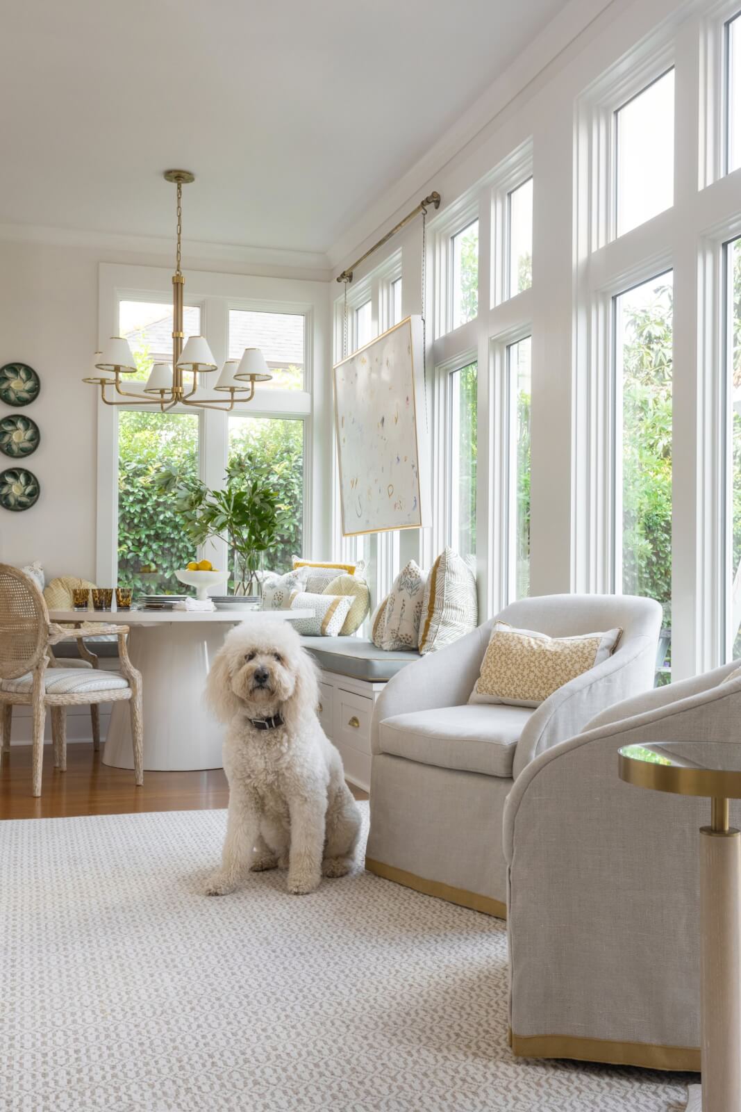 Bright room with large windows, beige furniture, and a round table surrounded by chairs. A fluffy white dog rests on a light carpet, enhancing the cozy home decor. Greenery is visible outside the windows, adding to this New Orleans home's natural charm.