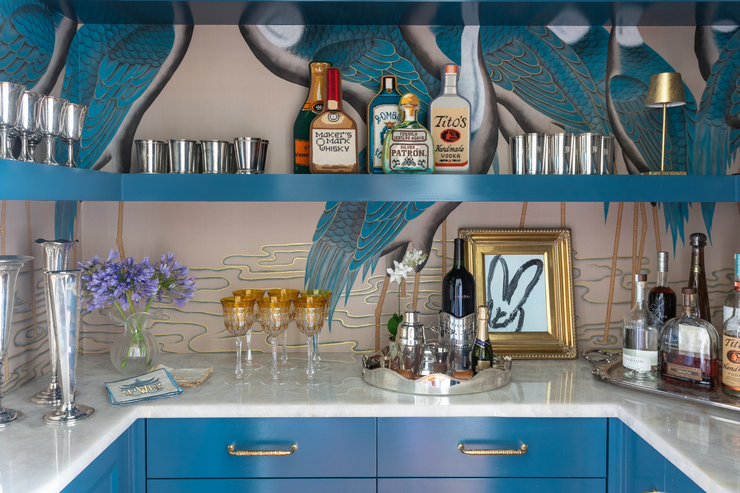 A New Orleans home bar featuring the prettiest wallpaper adorned with birds frames a collection of whiskey and vodka bottles. Metal cups and cocktail glasses elegantly sit on a blue shelf, accompanied by a framed picture in the background.