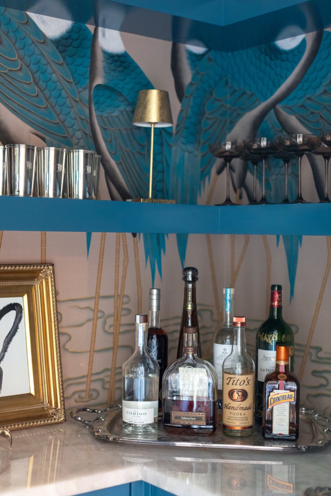 Bar shelf showcasing an assortment of liquor bottles, cocktail glasses, a framed picture, and a small lamp. The background boasts the prettiest wallpaper with an intricate blue and gold pattern that adds charm.