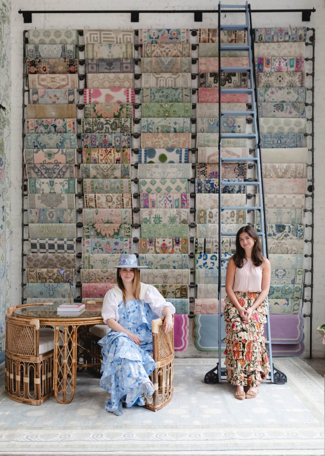 Two women posing in a bohemian-style interior with a wall of decorative tiles, sitting near a wooden table with wicker chairs.