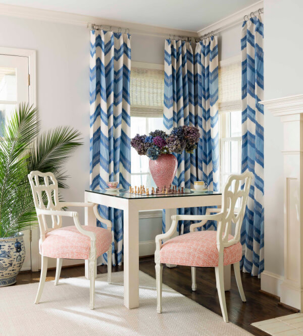 A small table with a chessboard and vase of flowers is flanked by two white wooden chairs with pink cushions, creating a charming feature in Roz Murphy's cozy home. Blue and white curtains frame the room, while a plant and fireplace add warmth in the background.