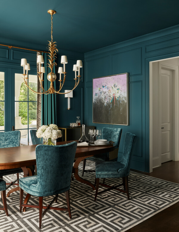 Embracing 2025 interior design trends, this dining room features teal walls and velvet chairs, anchored by a wooden table on a geometric rug. A chandelier adds elegance, while large windows flood the space with natural light. An abstract painting completes this harmonious blend of style and comfort.