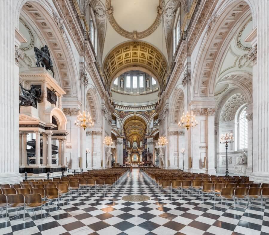 Interior of a grand cathedral in London, with ornate arches, a checkered floor, rows of wooden chairs, and decorative ceilings adorned with chandeliers.