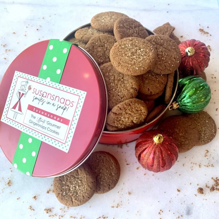 Open tin of ginger snap cookies with a vibrant pink and green lid, courtesy of one of the finest cookie companies that ship. Two small, decorative red and green ornaments are artfully arranged around the tin on a pristine white surface.