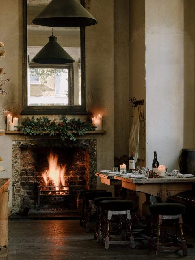 Cozy room with a lit fireplace, candles, and greenery on the mantel, perfect for a long weekend in London. A dining table set for meals awaits under large pendant lights hanging overhead.