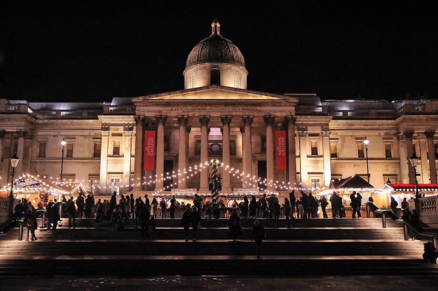 A neoclassical building with a grand dome stands illuminated against the night sky, its string lights twinkling like stars. Crowds bustle below, capturing the vibrant energy of a long weekend in London.