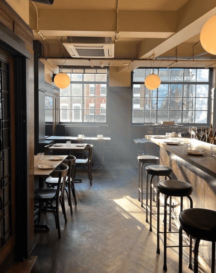 Sunlit cafe interior with wooden tables and chairs to the left, and a bar with stools to the right. Large windows allow natural light to fill the space—a perfect spot to unwind during a long weekend in London.