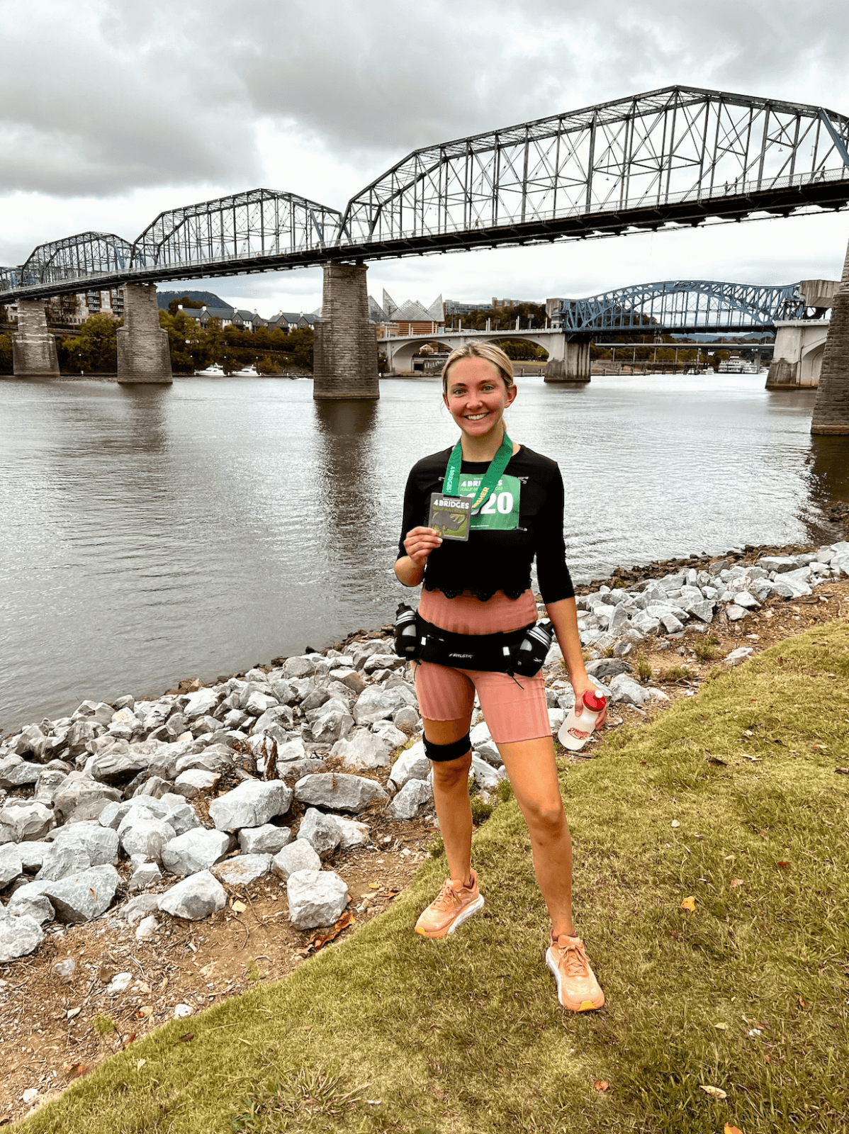 A person stands smiling by a river, holding a medal and drink bottle, with a bridge in the background—a perfect snapshot from one of the best spring marathons.