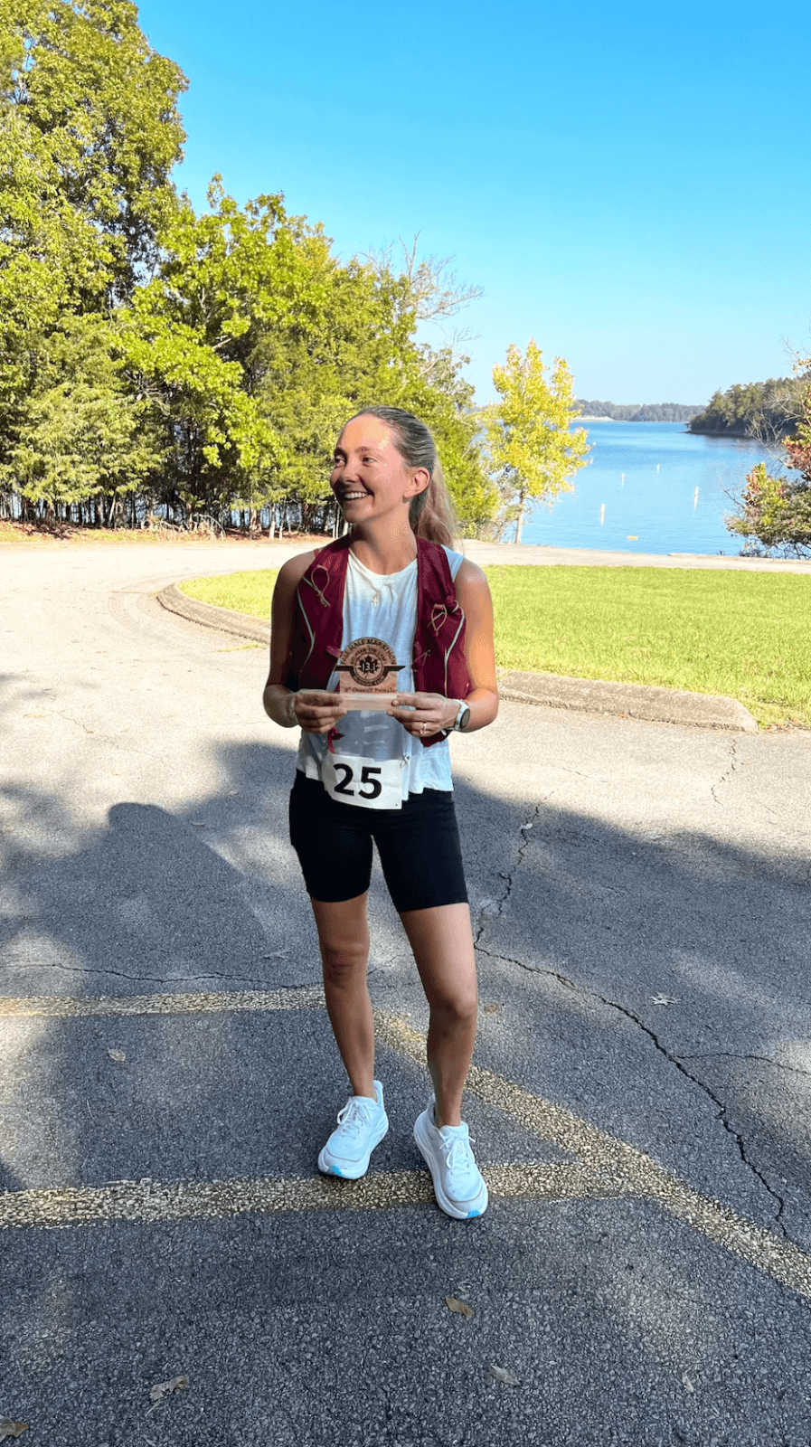 A woman stands on a paved path near a lake, holding a small plaque. She is dressed in athletic gear with the number 25 on her shorts. Trees and grass frame the scenic backdrop.