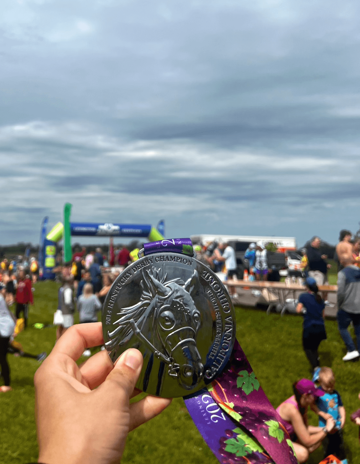 At an outdoor event bustling with people and framed by an inflatable arch, a person proudly holds a horse-themed race medal.