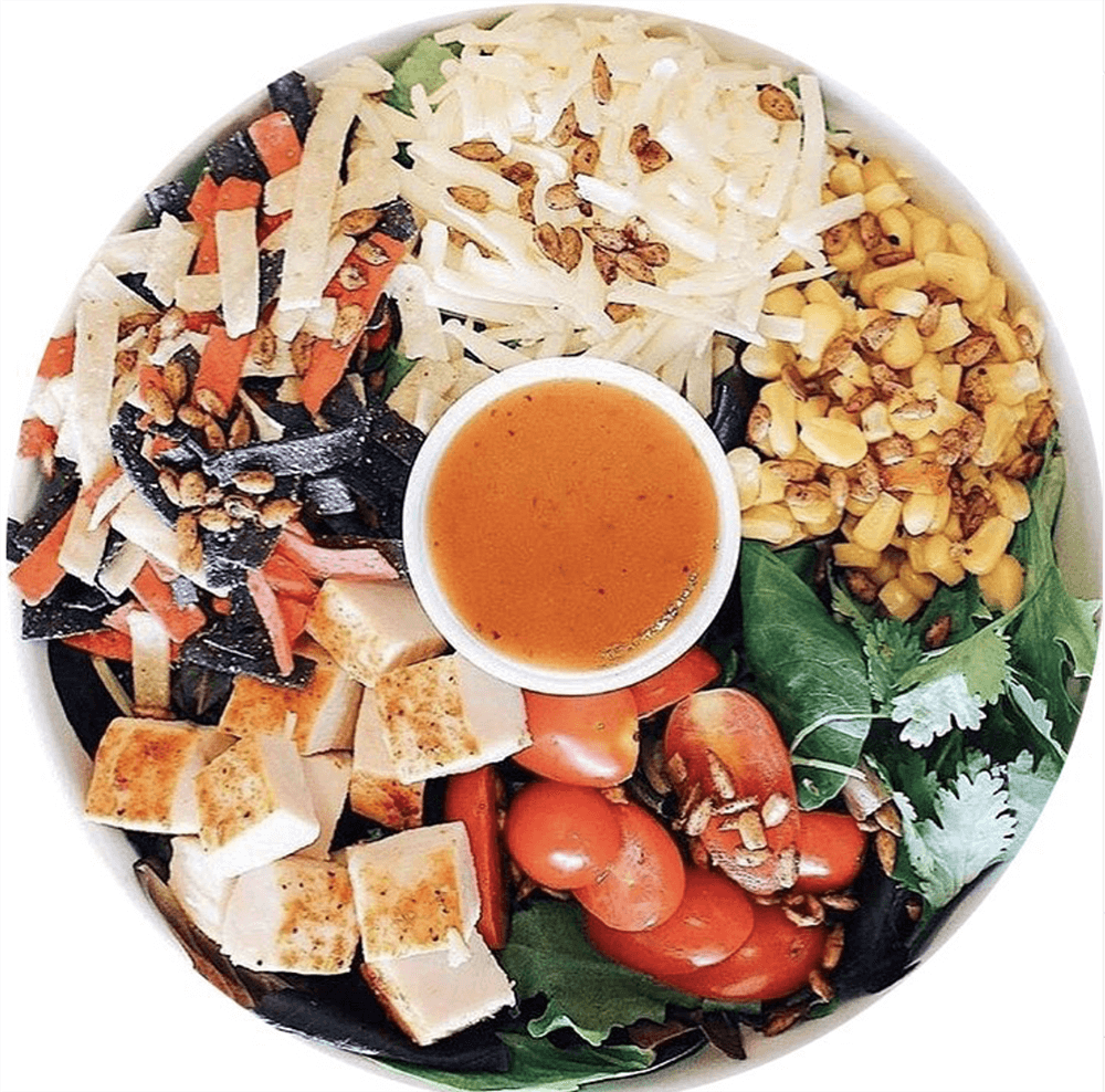 Discover one of Birmingham's best salads: a vibrant mix featuring grilled chicken, cherry tomatoes, mixed greens, corn, sliced vegetables, shredded cheese, seeds, and a cup of dressing in the center.