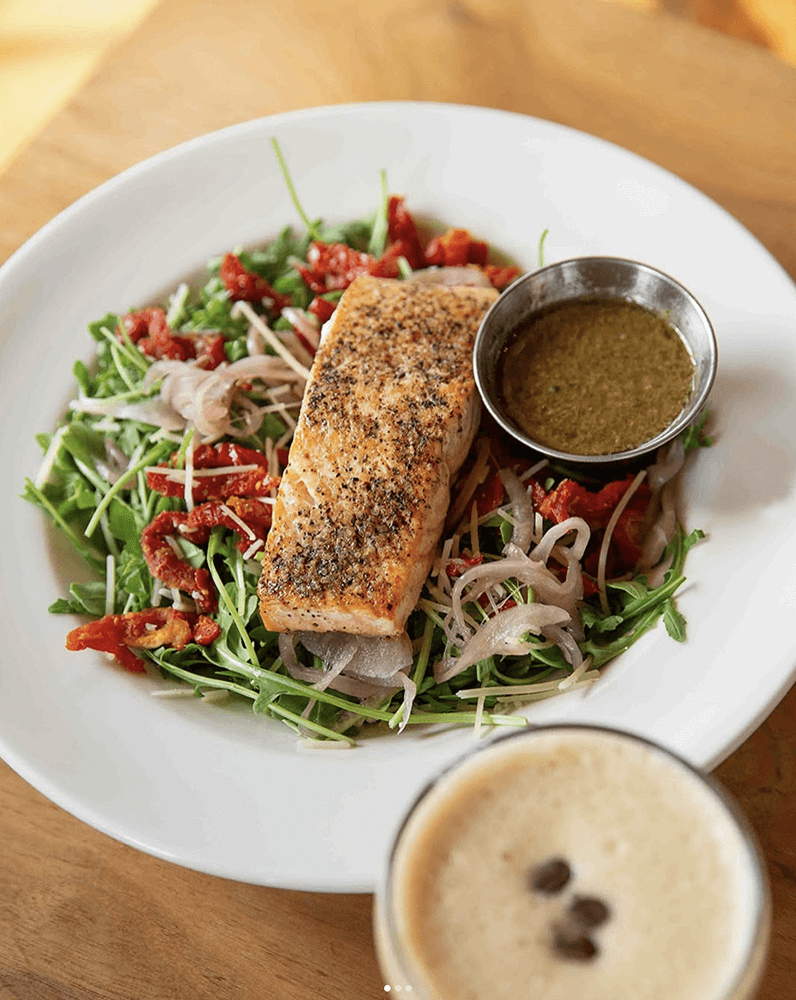 A plate of grilled salmon rests on a vibrant bed of arugula, shallots, and sundried tomatoes, capturing the essence of the best salads you'll find in Birmingham. A side of dressing complements this feast, while a steaming cup of coffee with beans on top completes the delightful scene.