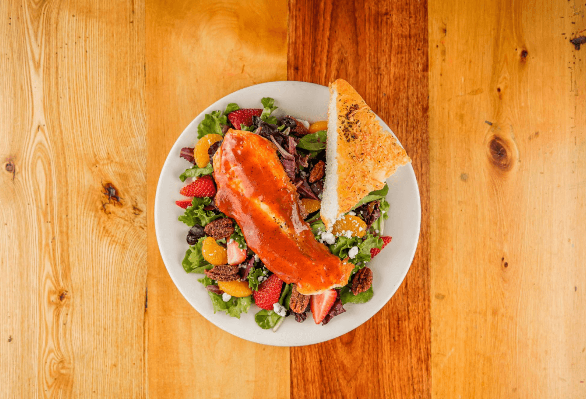 A top contender among the best salads in Birmingham, this delightful mix features crisp greens, juicy strawberries, oranges, pecans, and a protein on a white plate. It's perfectly paired with a slice of seasoned bread on a wooden table.