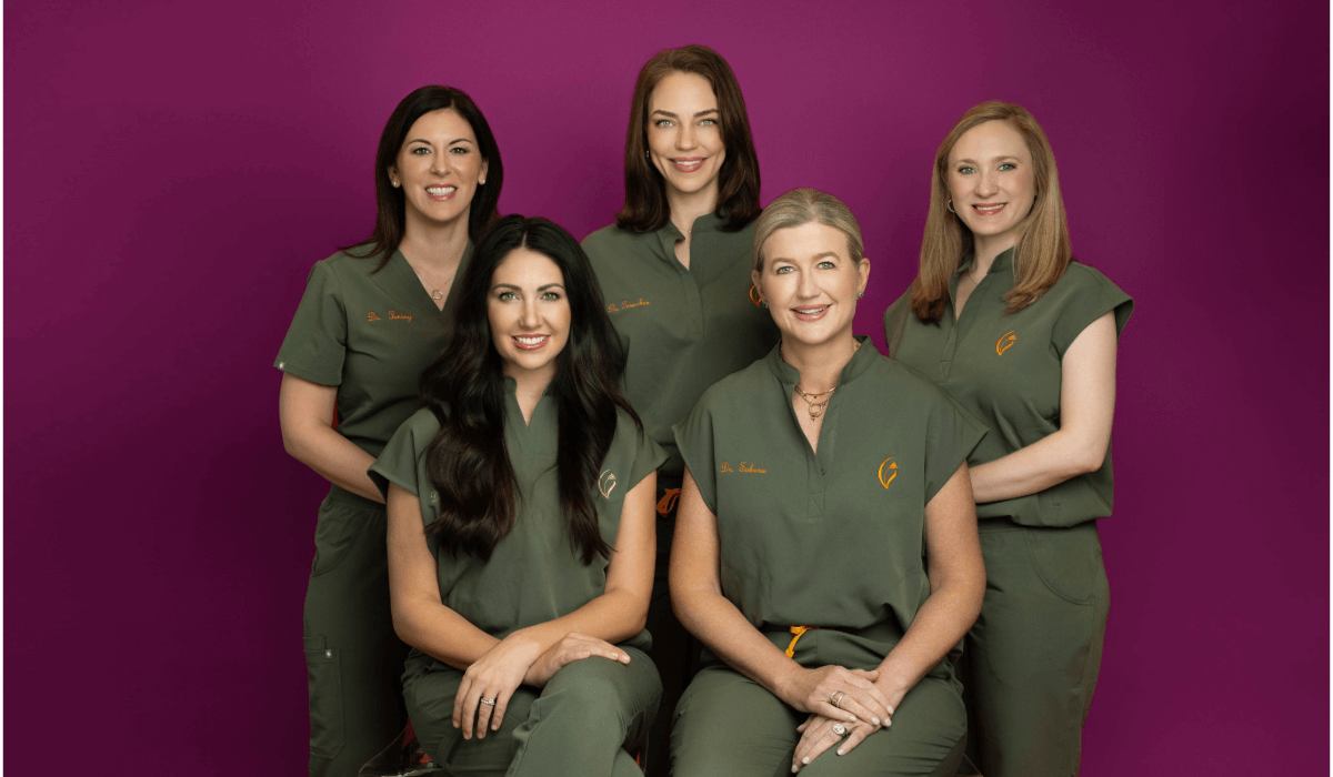 Five women in matching green scrubs pose in front of a purple background, representing the skilled team at Village Dermatology.