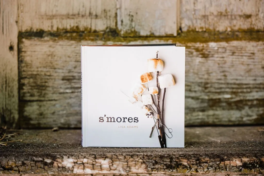 A cookbook titled "s'mores" by Lisa Adams, featuring toasted marshmallows on sticks, is propped against a weathered wooden background.