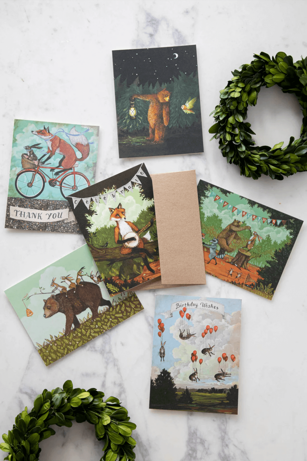 A selection of illustrated greeting cards featuring animals in whimsical scenes, like a fox cycling and a bear with balloons. Two green wreaths are placed near the cards on a marble surface.