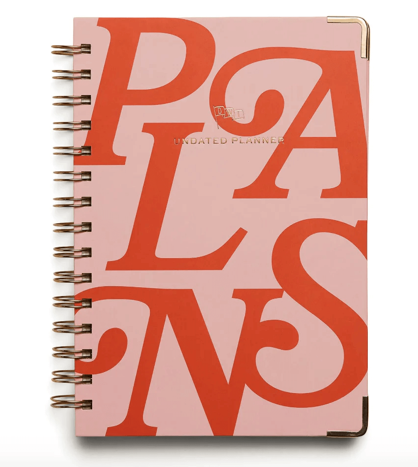 A spiral-bound planner with a pink cover and large orange letters spelling PLANS. It features gold corner accents and the words UNDATED PLANNER.