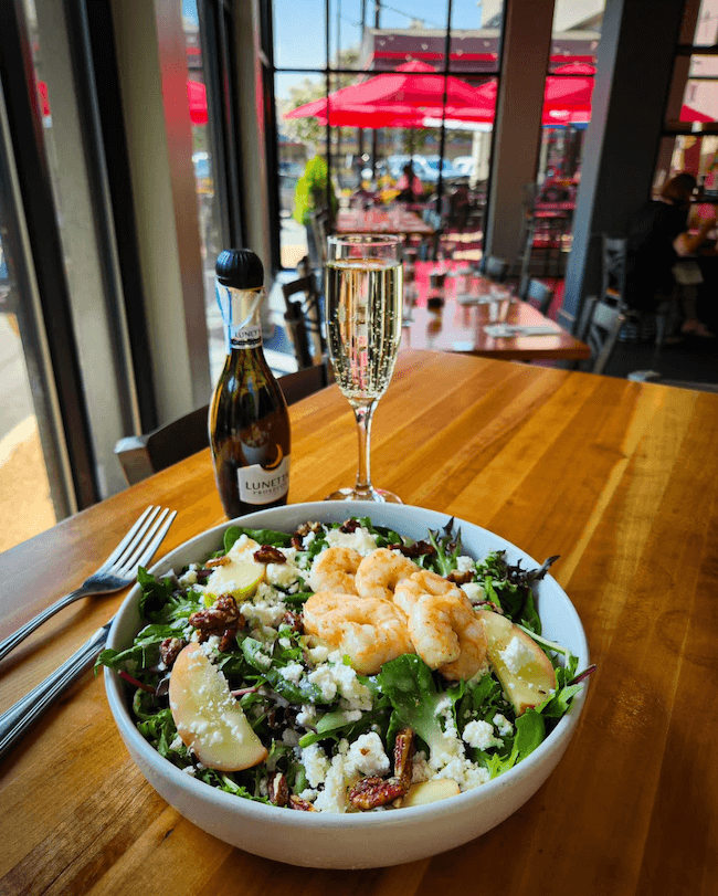 A bowl of salad with shrimp, apple slices, and cheese sits enticingly on a table next to a bottle and glass of sparkling wine—an experience worthy of being ranked among the best salads in Memphis.