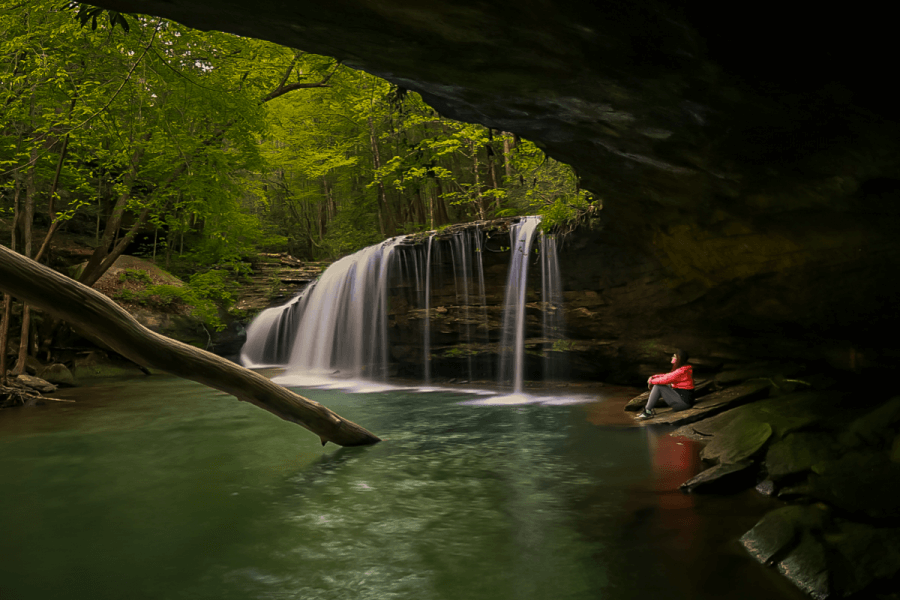 A person in a red jacket sits on a rock near a serene waterfall along the Waterfall Trail in the lush Kentucky Wildlands, with cascading water and a fallen tree in the foreground.