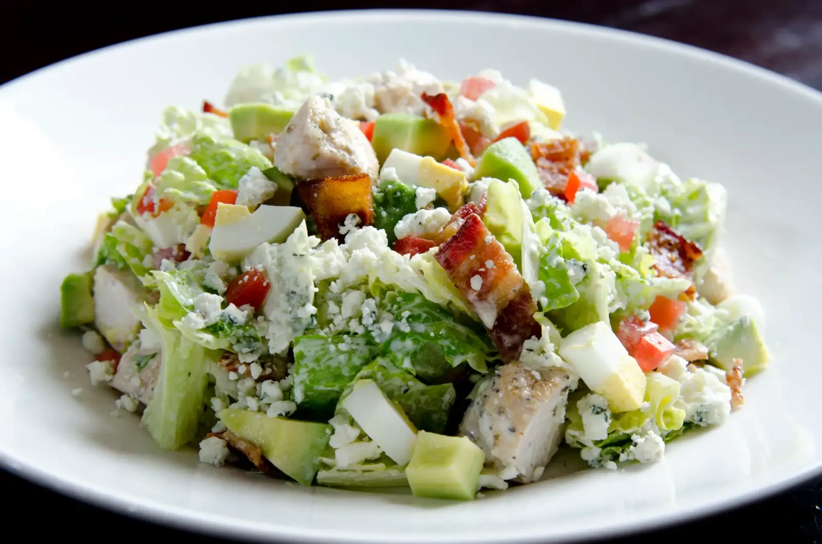 Discover one of Birmingham's best salads: a colorful Cobb with romaine lettuce, diced chicken, crispy bacon, egg, creamy avocado, juicy tomato, and tangy blue cheese elegantly arranged on a white plate.