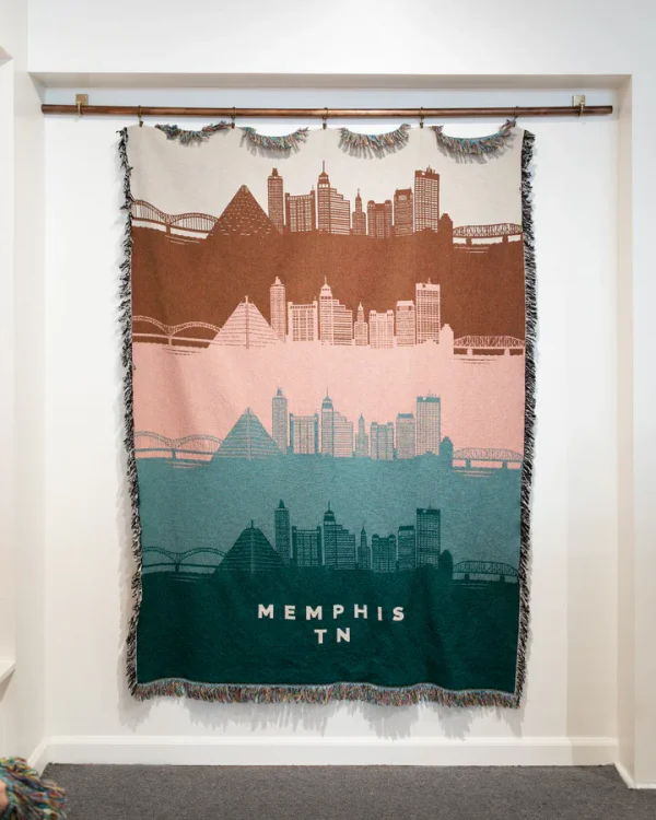 A woven tapestry elegantly captures the Memphis, TN skyline in brown, pink, and teal hues. Displaying iconic architectural landmarks with fringe edges, this stunning piece is hung on a rod in a white-walled room. Discover January 2025's finest artistic expression with Memphis FINDS.